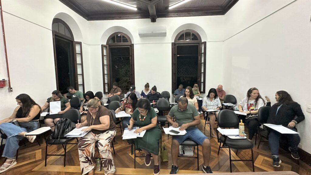 A imagem mostra uma sala de aula com várias pessoas sentadas em cadeiras, a maioria delas voltada para a frente, como se estivessem prestando atenção a uma aula ou palestra. Há janelas altas e arqueadas com venezianas de madeira, e o teto tem vigas de madeira aparentes. As paredes são brancas e a iluminação parece vir de luminárias no teto. As pessoas na sala estão em diferentes posições: algumas estão escrevendo em papéis sobre as mesas, outras parecem estar conversando entre si ou apenas observando. A maioria está vestida casualmente. O ambiente sugere um local de aprendizado ou treinamento. Há também um ar condicionado instalado na parede entre duas janelas.