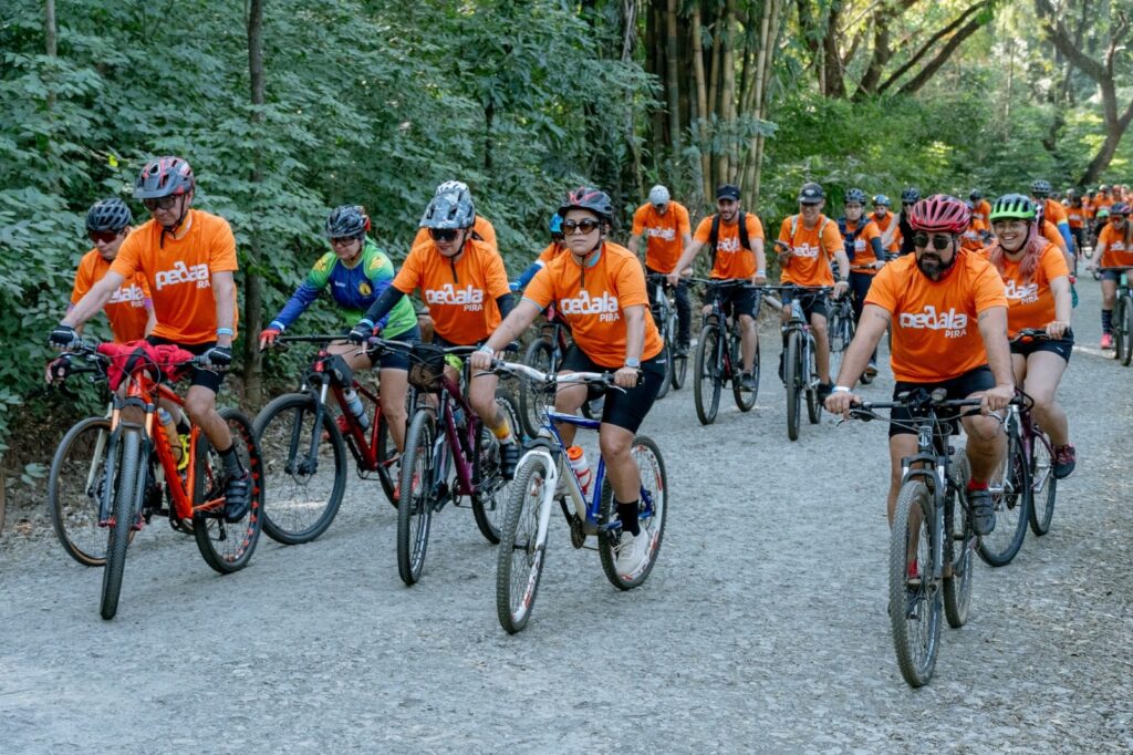 A imagem mostra um grupo de ciclistas pedalando em uma trilha de terra cercada por árvores e vegetação exuberante. A maioria dos ciclistas usa camisetas laranja vibrantes com o logo "pedala PIRA". Eles estão usando capacetes de ciclismo e óculos de sol, e suas bicicletas são mountain bikes. O dia parece ensolarado, com a luz do sol filtrando através das árvores. A cena transmite uma sensação de aventura e comunidade, com os ciclistas desfrutando de um passeio em meio à natureza.