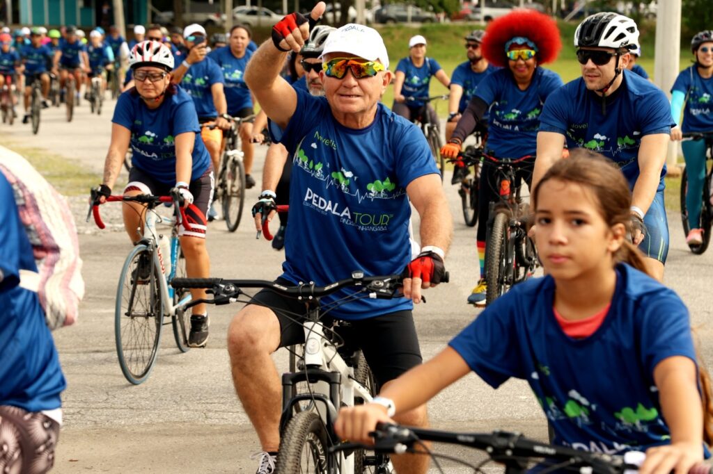 A imagem mostra um grupo de ciclistas participando de um evento, possivelmente um passeio ciclístico ou corrida. A maioria dos participantes está vestindo camisetas azuis com um logotipo que parece ser "PEDALA TOUR". No primeiro plano, um homem mais velho, com um boné branco e óculos de sol esportivos, acena para a câmera com a mão direita levantada, enquanto pedala. Uma menina, também vestindo uma camiseta azul, está em primeiro plano, olhando para frente. Vários outros ciclistas, incluindo mulheres e homens, podem ser vistos ao fundo, todos pedalando em uma estrada pavimentada. Há também uma mulher com um penteado afro vermelho vibrante, que se destaca no grupo. A atmosfera geral parece ser de um evento comunitário ou esportivo, com um clima animado.
