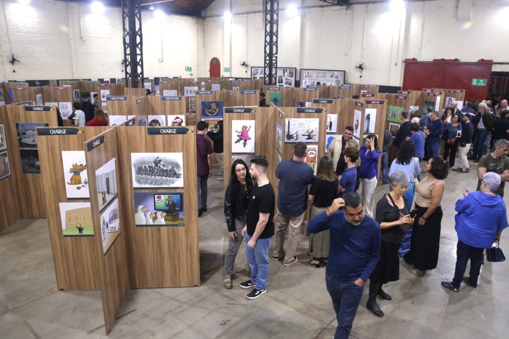 A imagem retrata uma exposição de charges, com diversas pessoas circulando entre os painéis de madeira que exibem as obras. Os painéis são organizados de forma a criar um labirinto, e em alguns deles, a palavra "CHARGE" está escrita em um suporte com formato de lápis. As charges expostas parecem abordar temas variados, com algumas apresentando desenhos de personagens e outras cenas mais complexas com figuras humanas. As pessoas na exposição estão vestidas de maneira casual e interagem entre si, observando as charges com interesse. A iluminação geral do espaço é artificial, vinda de luminárias no teto, e o chão é de cimento. Ao fundo, percebe-se a estrutura de um galpão industrial, com colunas metálicas e paredes de alvenaria.