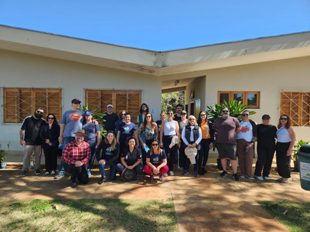 Um grupo de 25 pessoas, composto por 14 mulheres e 11 homens, está posando para uma foto em frente a um prédio com uma fachada de cor clara e janelas com ripas de madeira. O grupo está espalhado em duas fileiras, com algumas pessoas na frente agachadas. As pessoas estão vestidas de forma casual, e a atmosfera geral parece ser relaxada e amigável. O prédio tem uma área de entrada coberta, e há plantas e árvores visíveis ao fundo, sugerindo um ambiente externo. O céu está claro e azul, indicando um dia ensolarado.
