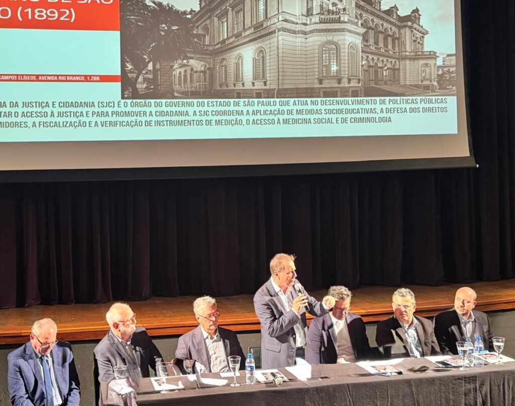 A imagem mostra um evento em um auditório, com uma tela projetando informações no fundo e seis homens sentados em uma mesa à frente. Na tela, há um texto que descreve o órgão "Justiça e Cidadania (SJC)", que é parte do governo do estado de São Paulo. Ele atua no desenvolvimento de políticas públicas, visando facilitar o acesso à justiça e promover a cidadania. A SJC também coordena a aplicação de medidas socioeducativas, a defesa dos direitos dos consumidores, a fiscalização e a verificação de instrumentos de medição, além de acesso à medicina social e criminologia. Há também uma imagem de um edifício em estilo clássico, possivelmente relacionado ao órgão mencionado. À frente da tela, uma mesa longa está coberta por uma toalha preta. Seis homens, todos de terno, estão sentados atrás da mesa. Um homem, posicionado levemente em pé e com um microfone na mão, parece estar falando para a plateia ou para os outros homens na mesa. Os outros cinco homens estão sentados, alguns com óculos, e parecem estar ouvindo atentamente. Objetos como garrafas de água, copos e papéis estão dispostos sobre a mesa. O ambiente sugere um evento formal, como uma palestra, seminário ou mesa redonda. A iluminação parece ser focada na área da mesa e do apresentador.