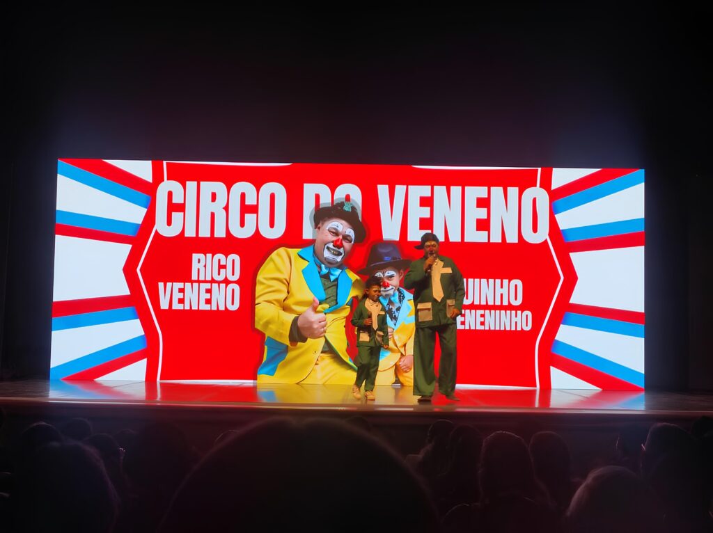 A imagem mostra um palco com uma tela de LED iluminada ao fundo, exibindo o título "CIRCO DO VENENO" em letras grandes e brancas sobre um fundo vermelho com detalhes em azul e branco nas laterais. No centro da tela, há imagens de três palhaços. Um palhaço maior, com um terno amarelo e gravata borboleta azul, está à esquerda, fazendo um sinal de "joinha" com o polegar. Ao lado dele, um palhaço menor, com um terno verde e chapéu, está em pé. À direita, um palhaço com um terno verde e um microfone na mão, também está em pé, aparentemente apresentando. Abaixo das imagens dos palhaços, estão escritos os nomes "RICO VENENO" à esquerda e "JUINHO ENENINHO" à direita. No palco, em frente à tela, estão os três palhaços que aparecem na imagem de fundo. Eles estão em poses semelhantes às das imagens projetadas. O público, cujas silhuetas são visíveis, está sentado na plateia escura, assistindo à apresentação. A iluminação do palco destaca os artistas e a tela.