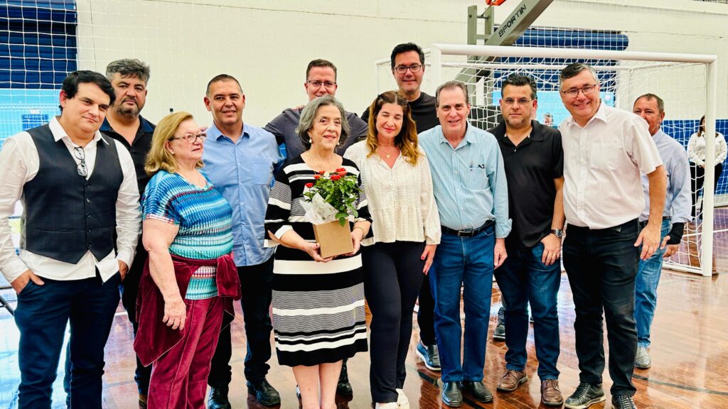 A imagem mostra um grupo de pessoas em um ginásio, possivelmente após uma competição esportiva. No centro, uma jovem atleta sorri, segurando um buquê de flores cor-de-rosa e uma medalha. Ela veste um agasalho esportivo nas cores azul e verde, com detalhes ondulados.À sua volta, homens posam para a foto. À esquerda, um homem com barba e cabelo grisalho veste uma camisa polo branca com um logo. Ao lado dele, outro homem, com camisa azul clara, sorri e parece segurar o buquê. Mais à direita, um homem de camisa xadrez azul aponta para a medalha da atleta, com um sorriso largo. Atrás deles, outros homens e uma mulher completam o grupo. O fundo exibe uma rede de proteção e a quadra do ginásio.