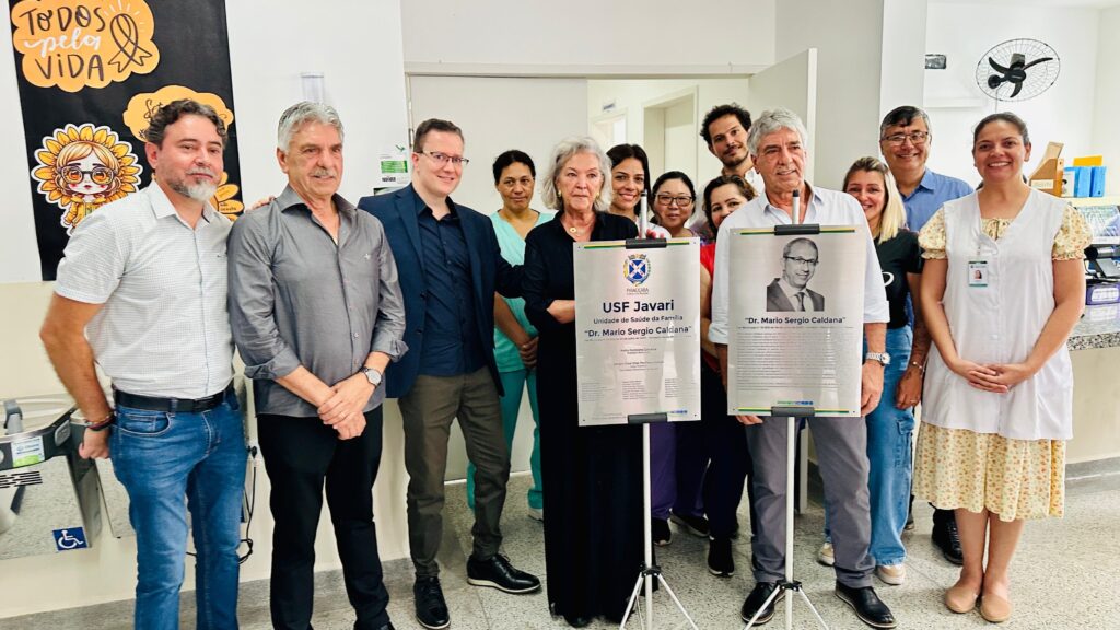 A imagem mostra um grupo de pessoas em um evento de inauguração ou homenagem em uma unidade de saúde. No centro, há uma placa com o nome "USF Javari" e "Dr. Mario Sergio Caldana", indicando que a unidade está sendo nomeada em homenagem a essa pessoa. As pessoas em volta parecem estar participando da cerimônia, com algumas delas posando para a foto ao lado da placa. Há também uma faixa decorativa com a frase "TODOS pela VIDA" e um desenho de um girassol. A atmosfera geral sugere um evento positivo e comunitário.