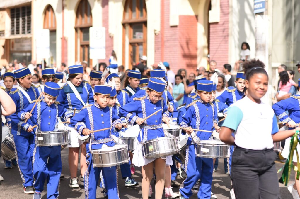 A imagem mostra um grupo de crianças e adolescentes vestindo uniformes de banda azul e dourada, marchando em uma rua. Eles estão tocando tambores com baquetas. Os uniformes incluem chapéus azuis com adornos dourados e penas brancas, jaquetas azuis com detalhes dourados e calças azuis. Alguns dos jovens estão olhando para a frente, enquanto outros parecem concentrados em tocar seus instrumentos. Ao fundo, há uma multidão de pessoas observando a marcha, e edifícios com janelas arqueadas. Uma menina em primeiro plano, à direita, está fora de foco e usa uma camiseta branca e preta. A atmosfera geral da imagem sugere um desfile ou evento cívico.