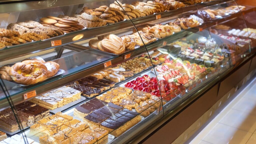 A imagem exibe uma vitrine de padaria repleta de uma variedade de doces e pães. As prateleiras estão organizadas com diferentes tipos de produtos, incluindo biscoitos, bolos, tortas e outros doces finos. Há uma seleção de biscoitos em destaque na prateleira superior, seguidos por pães doces, tortas de frutas e éclairs nas prateleiras inferiores. Os produtos são apresentados de forma atrativa, com iluminação que realça suas cores e texturas, convidando os clientes a escolherem suas guloseimas preferidas.