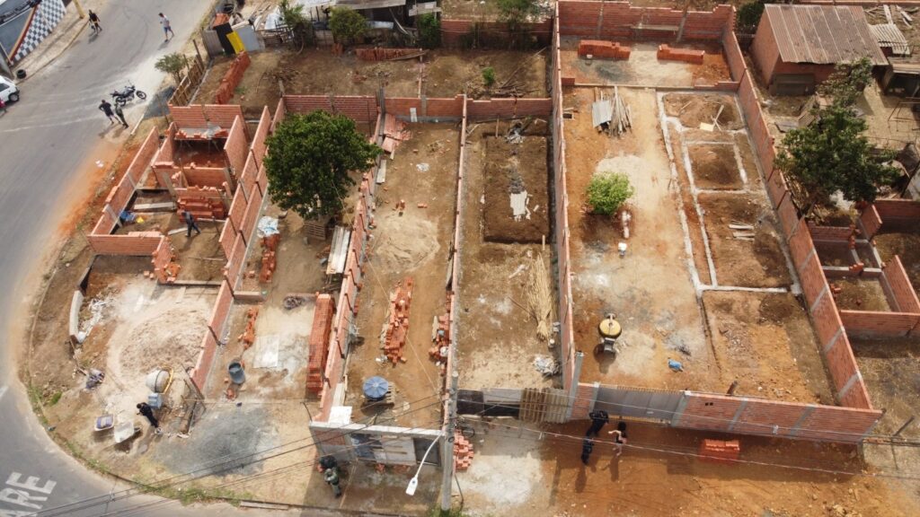 A imagem mostra uma construção em andamento, vista de cima. Várias paredes de tijolos estão em diferentes estágios de construção. Há pilhas de tijolos espalhadas pelo local, indicando material de construção. No chão, há sinais de trabalho, como uma betoneira e carrinhos de mão. Pessoas vestindo roupas de trabalho podem ser vistas em diferentes áreas do canteiro de obras, envolvidas em atividades de construção. O terreno é em grande parte de terra batida, com algumas áreas de vegetação e árvores. Ao redor do canteiro de obras, há uma rua com alguns veículos e pessoas, sugerindo um ambiente urbano ou suburbano. A construção parece ser de várias unidades residenciais ou comerciais.