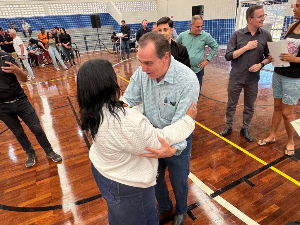 A imagem mostra um momento caloroso em um ginásio coberto, com piso de madeira polida e uma parede com rede ao fundo. Em destaque, um homem de camisa azul clara e jeans cumprimenta ou abraça afetuosamente uma mulher vestida com jaqueta branca acolchoada e calça azul, transmitindo um clima de cordialidade e respeito. Ao redor, outras pessoas estão em pé ou sentadas, algumas segurando documentos.