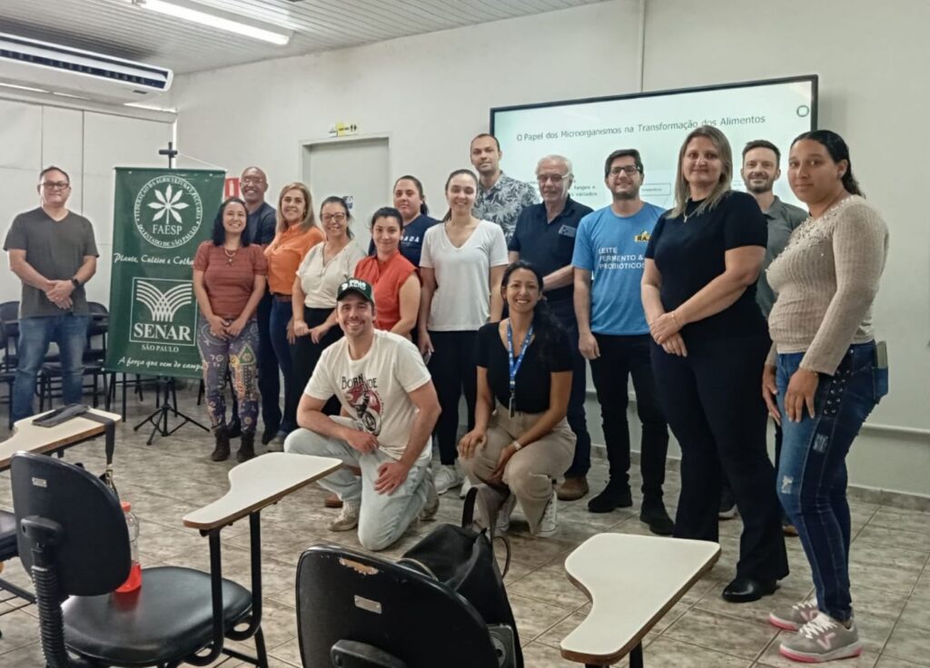 A imagem mostra um grupo de 16 pessoas em uma sala de aula, possivelmente após uma apresentação ou treinamento. No fundo, há um banner verde com o logo do SENAR (Serviço Nacional de Aprendizagem Rural) e o slogan "Plante, Cultive & Colha. A força que vem de campo." Acima do banner, há uma projeção em uma tela com o título "O Papel dos Microorganismos na Transformação dos Alimentos" e alguns tópicos secundários. As pessoas estão vestidas de forma casual e variada, com algumas delas em pé e outras agachadas na frente. Há mesas e cadeiras dispostas na sala, sugerindo um ambiente de aprendizado. A iluminação é artificial, vinda de cima, e a atmosfera parece ser de um evento de formação ou workshop.