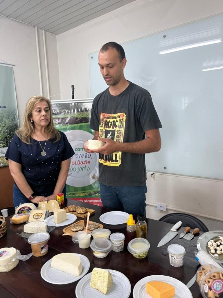 A imagem mostra duas pessoas em pé atrás de uma mesa cheia de queijos e acompanhamentos. Um homem, vestindo uma camiseta do AC/DC, segura um pedaço de queijo branco em sua mão e parece estar explicando algo. Ao lado dele, uma mulher com cabelo loiro e uma blusa azul escura observa atentamente. A mesa está repleta de uma variedade de queijos, alguns em embalagens, outros já cortados em diferentes formas e tamanhos. Há também potes com molhos ou pastas, mel, azeite e utensílios para cortar e servir queijo, como facas e espátulas. Ao fundo, um banner com imagens verdes e texto em português sugere que o evento está relacionado à agricultura ou produção de alimentos.