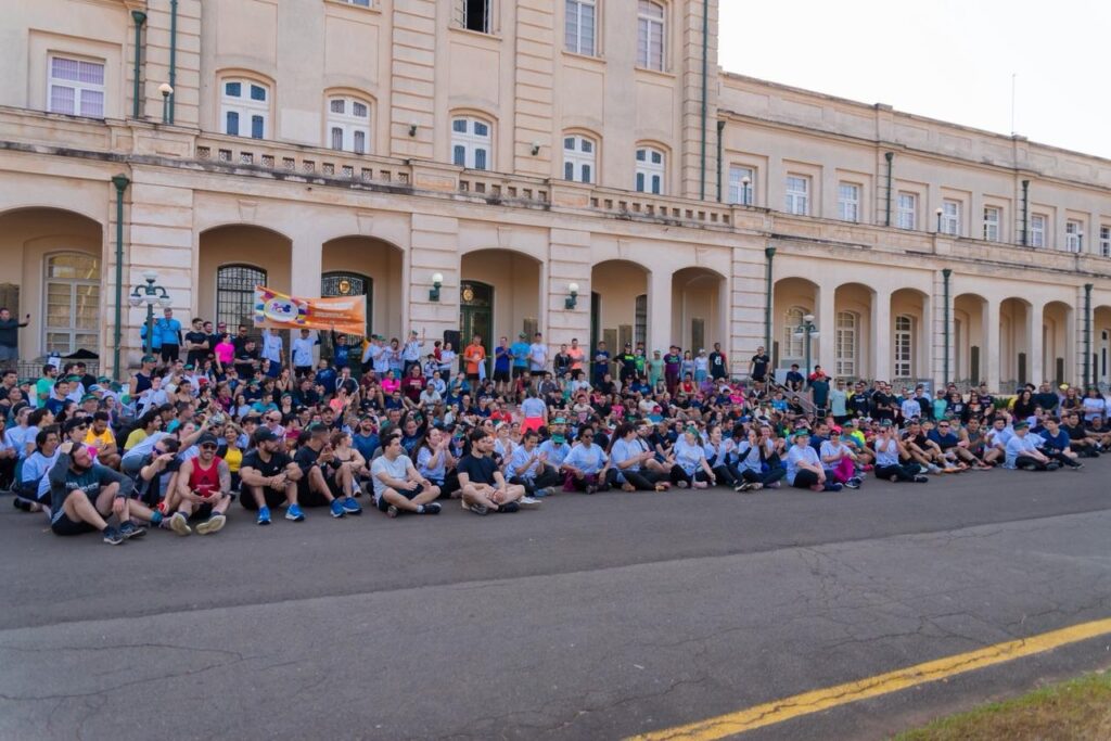 A imagem mostra um grande grupo de pessoas sentadas no chão, em frente a um edifício histórico com colunas e arcos. A maioria das pessoas está vestida casualmente, em cores variadas, e parece estar participando de um evento ou reunião ao ar livre. No centro da multidão, há uma faixa laranja com desenhos e texto, que sugere que o evento pode estar relacionado a alguma causa ou organização. Algumas pessoas estão em pé atrás da multidão sentada, e atrás delas, a fachada do edifício se estende amplamente, com várias janelas e detalhes arquitetônicos. O céu está claro, indicando um dia ensolarado.