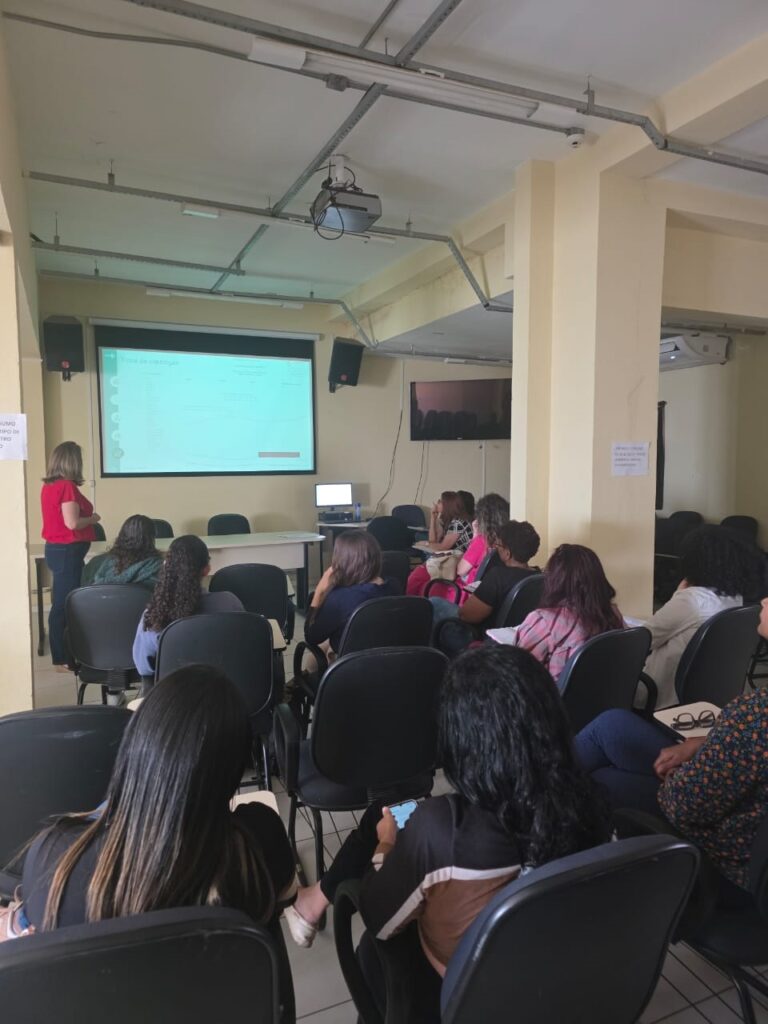 A imagem mostra uma sala de aula ou auditório com um grupo de pessoas sentadas em cadeiras, assistindo a uma apresentação projetada em uma tela. Uma mulher, vestida com uma blusa vermelha, está de pé perto da tela, possivelmente apresentando ou instruindo o grupo. Há um projetor no teto e caixas de som nas laterais da tela. A iluminação é artificial, vindo de luminárias no teto. O ambiente parece ser um local de aprendizado ou reunião.