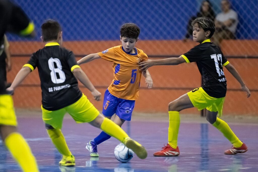 A imagem mostra uma partida de futsal entre jovens jogadores. No centro, um jogador com a camisa laranja e o número 5 tenta controlar a bola, cercado por dois adversários. Um deles, vestindo a camisa preta com o número 8 e meias amarelas, se aproxima pela esquerda, enquanto o outro, com a camisa preta e o número 13 e meias amarelas, avança pela direita. O jogador de laranja está em movimento, com a bola aos seus pés, e parece estar em uma disputa pela posse. O fundo da imagem exibe a quadra com suas marcações e uma grade de proteção, com algumas pessoas assistindo ao jogo ao fundo. A iluminação é clara, destacando a ação dos jogadores.