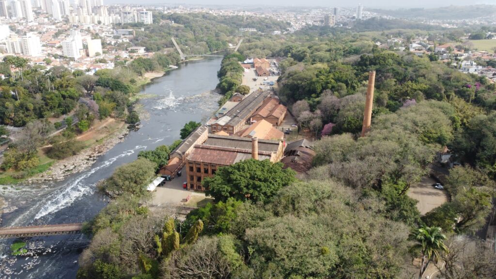 A imagem é uma vista aérea de uma paisagem urbana e natural, com destaque para um rio que atravessa a cena. À esquerda, um rio com corredeiras e uma ponte metálica sobre ele. Ao longo da margem do rio, há uma área verde com árvores e vegetação densa. No centro da imagem, um complexo industrial com edifícios de tijolos aparentes e telhados de cor alaranjada, com uma chaminé de tijolos alta e imponente. O complexo parece ser antigo, com um estilo arquitetônico industrial clássico. À direita e ao fundo, uma cidade com prédios altos e modernos, contrastando com a arquitetura histórica do complexo industrial. A paisagem é dominada por uma grande quantidade de árvores e vegetação, que se estendem por toda a área, criando um ambiente de transição entre a natureza e a urbanização. A luz do sol ilumina a cena, criando sombras e realçando as texturas da água, das construções e da vegetação.