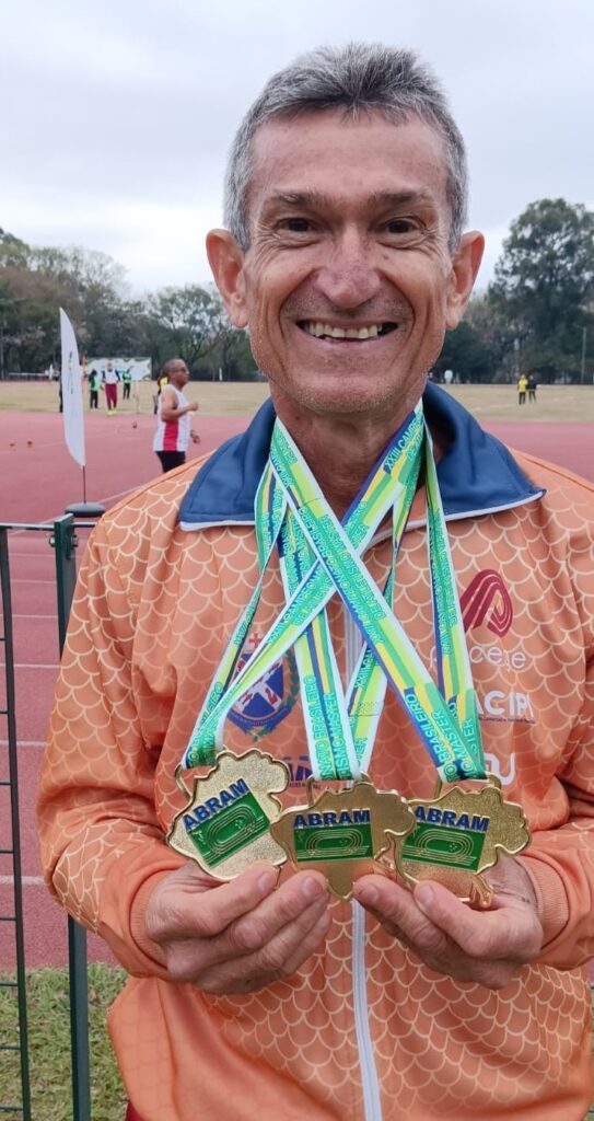 O homem na foto está sorrindo para a câmera, segurando três medalhas douradas. Ele está usando uma jaqueta laranja com um padrão de escamas e várias medalhas penduradas em seu pescoço. As medalhas têm fitas verdes, amarelas e azuis com texto escrito nelas. O fundo da foto mostra uma pista de atletismo com algumas pessoas ao longe.