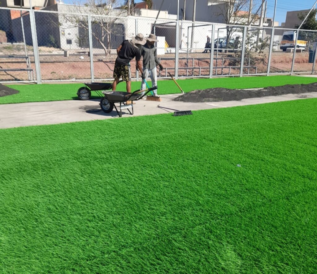 A imagem mostra dois trabalhadores instalando grama sintética em um campo. Eles estão usando chapéus para se proteger do sol e carregando materiais em carrinhos de mão. Há pilhas de areia ou borracha no chão, que são usadas para preencher a grama sintética. O campo está quase todo coberto com a grama verde vibrante. Ao fundo, há uma cerca e alguns edifícios, sugerindo que o campo está localizado em uma área urbana ou suburbana.