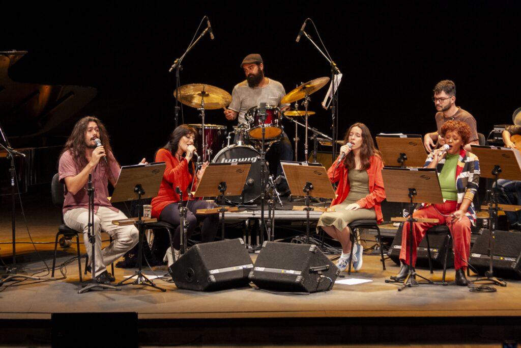 A imagem retrata um grupo de músicos se apresentando em um palco. No centro, um baterista com barba e boné toca com um conjunto de bateria. À frente dele, quatro vocalistas estão sentados em cadeiras, cada um com um microfone e um atril com partituras. Os vocalistas estão vestidos em cores variadas: um homem com cabelos longos usa uma camisa rosa e calças claras; uma mulher ao seu lado veste um suéter laranja vibrante; outra mulher, à direita, usa um blazer laranja sobre um vestido verde; e por último, uma mulher com cabelo cacheado vestida com uma jaqueta colorida, blusa verde e calças vermelhas. O palco é escuro ao fundo, com iluminação focada nos artistas. Há equipamentos de som e microfones espalhados pelo palco, indicando uma performance musical profissional. Ao fundo, à esquerda, parte de um piano é visível, sugerindo a presença de outros instrumentos na banda.