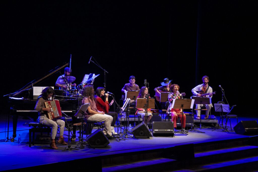 A imagem apresenta um grupo de músicos em um palco com fundo preto. Um piano de cauda está do lado esquerdo do palco, com um músico sentado tocando. À direita do piano, um baterista é visível atrás de um set de bateria. No centro do palco, vários músicos estão sentados, cada um com um suporte de música à sua frente. Um músico está tocando um acordeão, enquanto outros parecem estar cantando ou tocando outros instrumentos. A iluminação no palco é predominantemente azul, lançando um brilho vibrante sobre os performers e os instrumentos. A atmosfera geral sugere uma apresentação musical ao vivo.