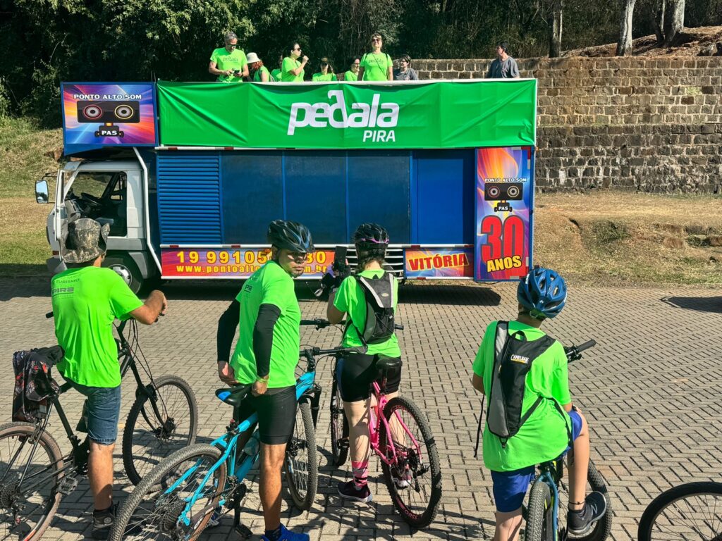 A imagem mostra um grupo de ciclistas, a maioria vestindo camisetas verde-limão, reunidos em torno de um caminhão. O caminhão tem uma grande faixa verde na frente com as palavras "pedala PIRA" escritas em branco. Há também um logotipo com "PONTO ALTO SOM" e um desenho de alto-falantes. Na lateral do caminhão, há um número de telefone (19 99105 302) e um site (www.pontoalto...). Há também uma inscrição "VITÓRIA 30 ANOS" com um grande número "30" em vermelho. As pessoas estão em um ambiente externo, com árvores e um muro de pedra visíveis ao fundo. Alguns dos ciclistas estão em suas bicicletas, enquanto outros estão em pé conversando. O dia parece ensolarado.