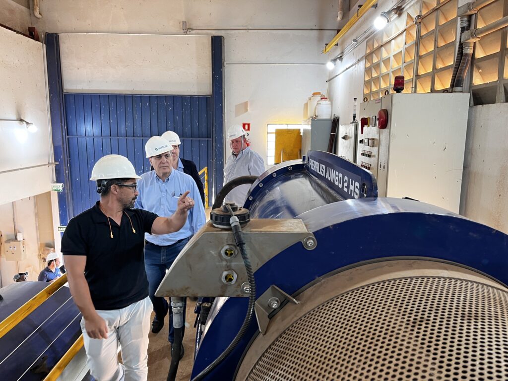 Um homem de camisa preta e calça branca, usando capacete branco e óculos, aponta para uma grande máquina azul com a inscrição "PIERLISI JUMBO 2 HS". Ele está em primeiro plano, de perfil, olhando para a máquina. Atrás dele, três homens de capacete branco observam a cena. Um deles, de camisa azul clara, também aponta para a máquina. Ao fundo, há uma porta azul grande, uma janela amarela e painéis elétricos. O ambiente parece ser industrial.
