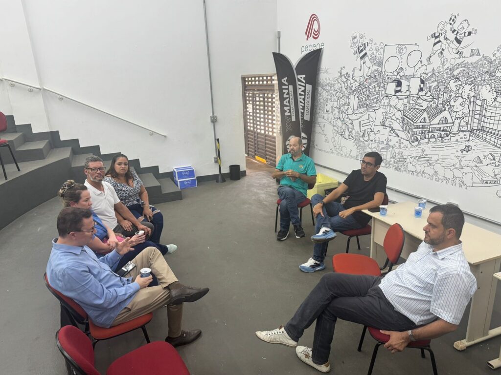 A imagem mostra um grupo de pessoas sentadas em cadeiras dispostas em semicírculo em uma sala. Há uma parede branca ao fundo, com uma escada à esquerda e uma parede decorada com um desenho em preto e branco à direita. No centro da sala, há duas bandeiras pretas com o texto "XV MANIA LOJA OFICIAL" em branco e vermelho. Há também um logo vermelho com o nome "pecene" no topo da parede decorada. As pessoas parecem estar em uma reunião ou discussão. Algumas estão segurando copos. A iluminação da sala é clara, vindo de cima.