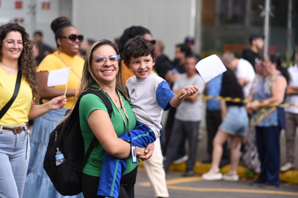 A imagem mostra um grupo de pessoas em um evento ao ar livre, possivelmente uma celebração ou manifestação. No centro, uma mulher com uma camiseta verde e óculos carrega um menino nos braços. Ambos sorriem para a câmera. O menino segura uma pequena bandeira branca em um palito. Ao lado da mulher, outra mulher com uma camiseta amarela e óculos escuros também sorri, segurando outra bandeira branca. Há outras pessoas ao fundo, algumas desfocadas, sugerindo que o evento está em andamento e há um fluxo de pessoas. A atmosfera parece animada e festiva.
