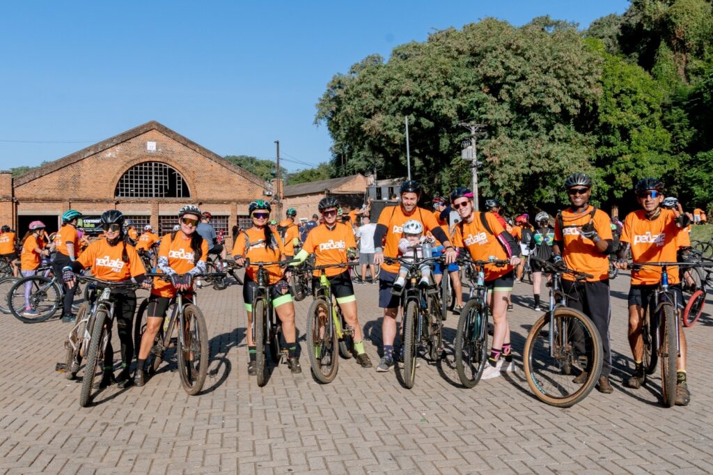 Um grupo de ciclistas, a maioria vestindo camisetas laranja com a inscrição "Pedala Pira", posa para uma foto em frente a um edifício de tijolos com um grande arco na fachada. Eles estão em uma área pavimentada com blocos de concreto, e há árvores verdes e um céu azul claro ao fundo. Os ciclistas estão em suas bicicletas, e todos usam capacetes. Alguns deles estão sorrindo para a câmera. Uma mulher no centro está segurando um bebê em uma cadeirinha de bicicleta na frente de sua bicicleta. A atmosfera geral é de um evento ao ar livre e comunitário.