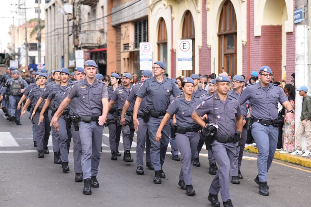 Diversos policiais militares desfilam em evento cívico por uma rua. Vestidos com uniforme cinza e boina cinza, homens e mulheres marcham, sendo que alguns olham para o público