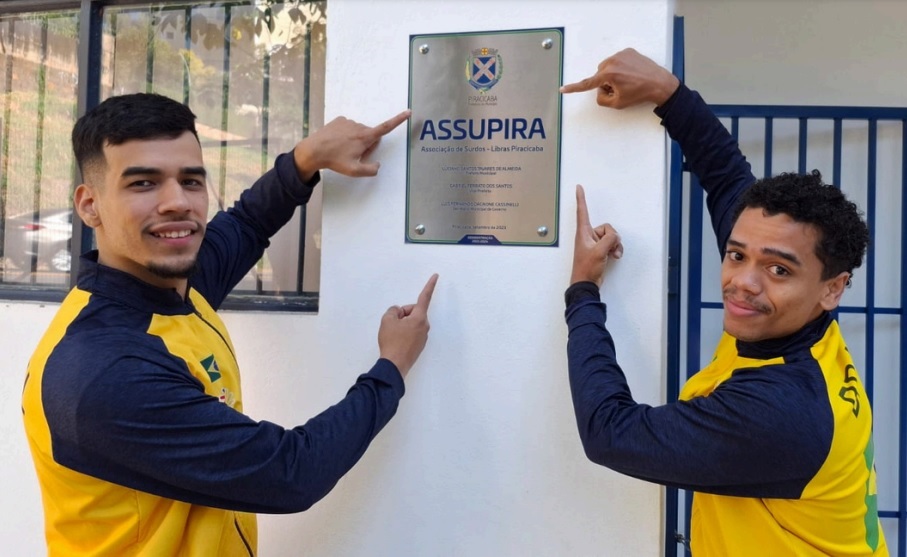 A imagem mostra dois jovens apontando para uma placa de inauguração. Ambos vestem jaquetas amarelas com detalhes em azul marinho. A placa, fixada em uma parede branca, exibe o brasão de Piracicaba e o nome "ASSUPIRA - Associação de Surdos - Libras Piracicaba". Abaixo, há nomes de pessoas e cargos, como "Gustavo Santos Tuarete de Almeida" e "Luiz Fernando Cardone Cassanello", além da data "24 de setembro de 2023". Um dos jovens tem pele clara e cabelo curto e escuro, enquanto o outro tem pele morena e cabelo encaracolado. Ao fundo, observa-se uma janela com grades e uma estrutura semelhante a um portão azul.