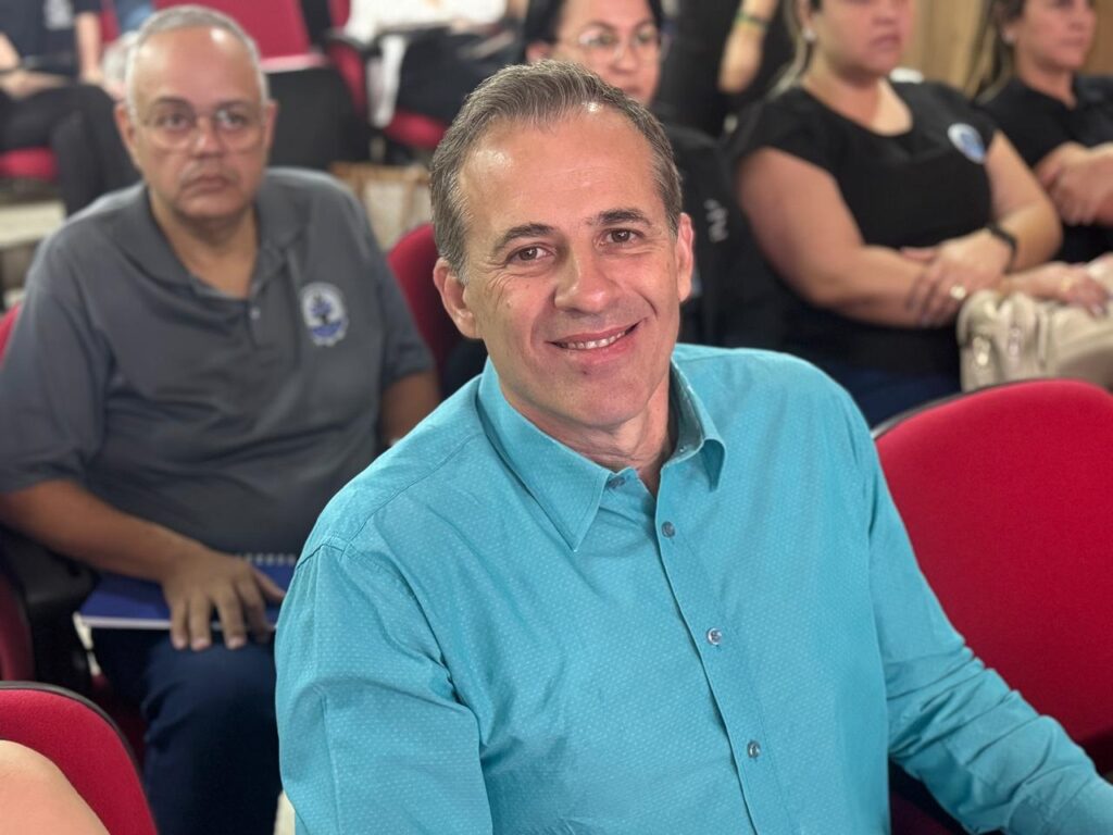 A imagem mostra um homem sorridente, vestindo uma camisa azul-clara com um padrão discreto, sentado em uma cadeira vermelha. Ele está em primeiro plano, e seu rosto é o foco principal. Ao fundo, outras pessoas estão sentadas em cadeiras, com algumas desfocadas. A iluminação parece ser artificial, vinda de cima, e cria algumas sombras no rosto do homem.