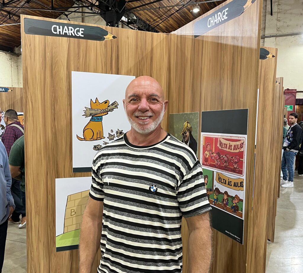 Jorge Lorde, sorridente, usa camiseta listrada em preto e branco e posa em frente a painéis de charges expostos no Salão Internacional de Humor de Piracicaba.