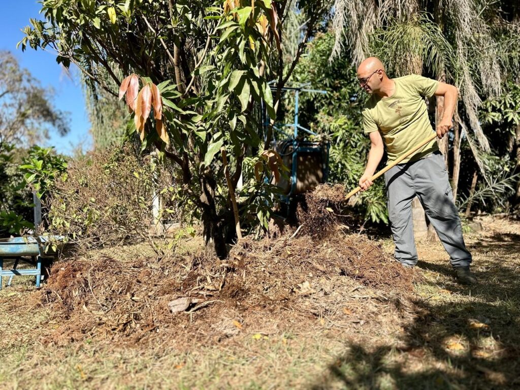 A imagem mostra um homem careca com óculos, vestindo uma camiseta verde e calças cinzas, usando uma pá para mexer um monte de terra e raízes em um jardim ensolarado. Ele está perto de uma árvore com folhas verdes e marrons. Ao fundo, há mais vegetação, palmeiras e um céu azul claro. A grama ao redor está seca e há uma estrutura azul metálica à esquerda.