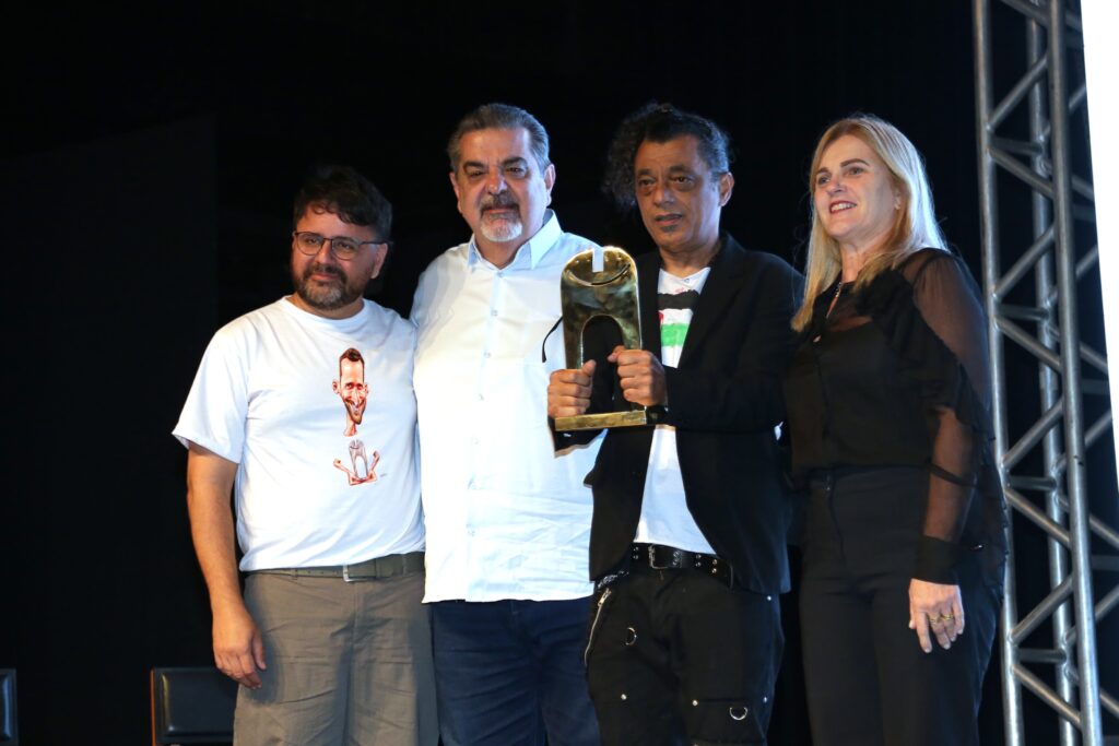 Quatro pessoas posam juntas no palco, Rui Miranda segura o troféu do Grande Prêmio.