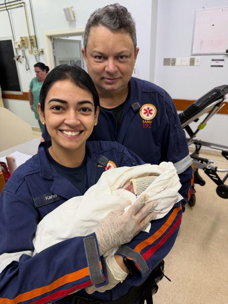 Na imagem, duas pessoas vestindo uniformes azuis do SAMU seguram um recém-nascido. A mulher, à esquerda, sorri para a câmera e segura o bebê enrolado em uma manta branca. Ela tem uma etiqueta com o nome "Karen" em seu uniforme. O homem, à direita, também sorri. Ambos parecem estar em um ambiente hospitalar, com equipamentos médicos e uma maca visíveis ao fundo. Uma outra mulher, vestida de verde, aparece ao fundo, de costas.