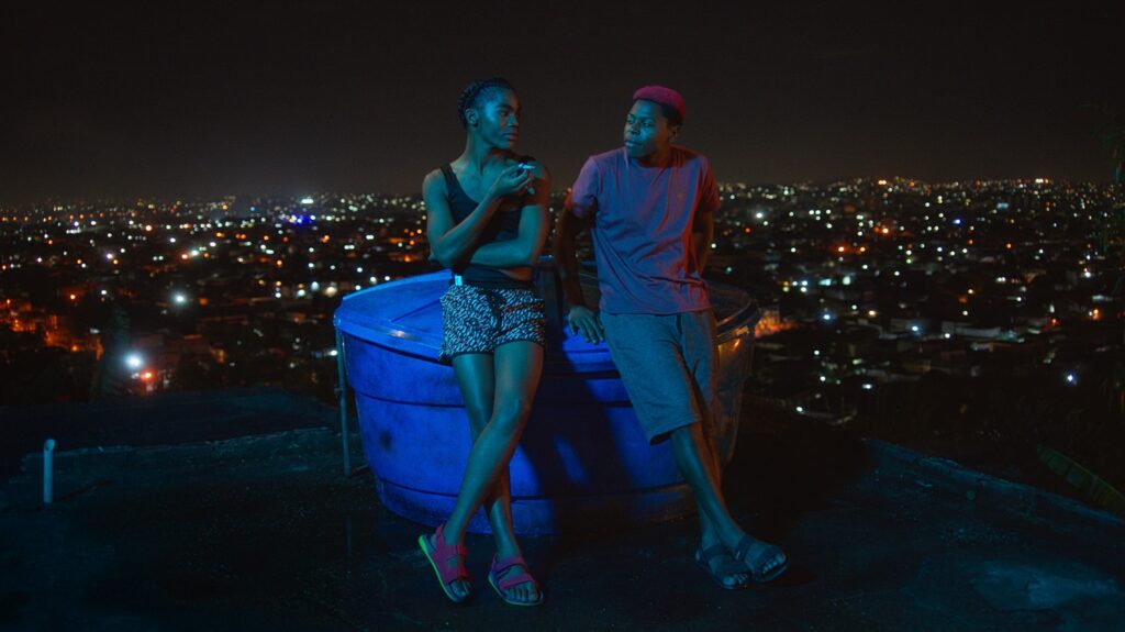 A imagem retrata dois jovens em pé, apoiados em um grande recipiente azul, à noite, com uma paisagem urbana iluminada ao fundo. A iluminação artificial cria um ambiente noturno, com luzes de casas e postes cintilando na cidade. Os jovens vestem roupas casuais e ambos parecem estar conversando. O jovem da esquerda usa um top preto e shorts estampados, segurando um cigarro. O jovem da direita usa uma camiseta vinho e shorts cinza, com um boné vermelho. A perspectiva da imagem sugere que eles estão em um ponto elevado, observando a cidade abaixo. A atmosfera geral é íntima e contemplativa.