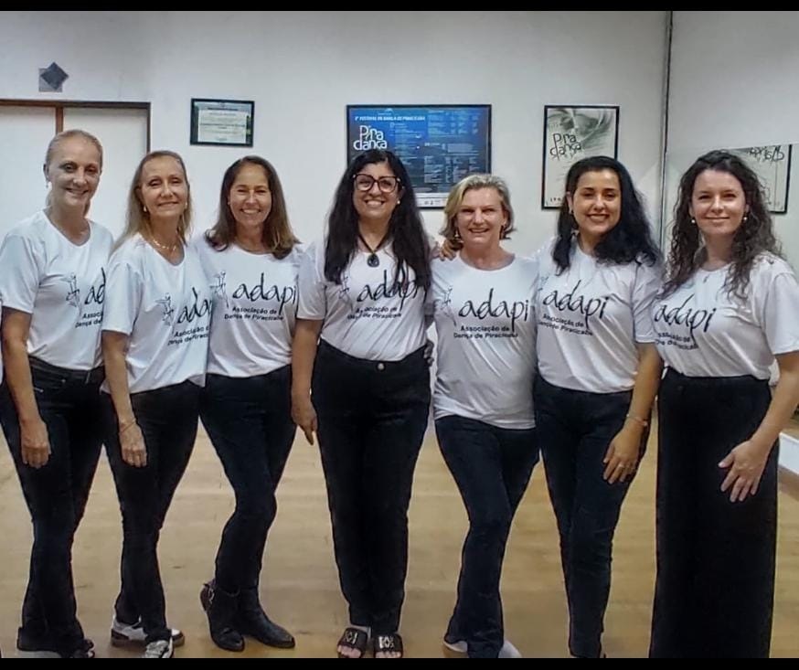 A imagem mostra um grupo de sete mulheres em pé, posando para uma foto. Todas estão vestindo camisetas brancas com um logotipo que diz "adapi Associação de Dança de Piracicaba" e um desenho estilizado de uma bailarina. As camisetas são combinadas com calças pretas de diferentes estilos. As mulheres estão alinhadas lado a lado, com sorrisos no rosto. A iluminação é suave e parece ser de um ambiente interno, possivelmente uma sala de dança ou estúdio, com espelhos refletindo ao fundo. Há quadros e pôsteres na parede atrás delas, um deles com o título "Pira Dança". A composição da foto é frontal, com as mulheres ocupando a maior parte do quadro.