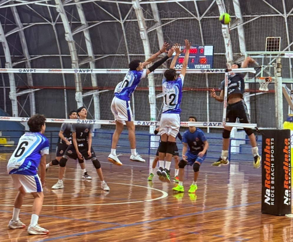 A imagem mostra uma partida de voleibol em quadra coberta. Os jogadores estão em ação, com alguns saltando para bloquear ou atacar a bola, que está no ar. A rede de voleibol divide a quadra. Há uma placa de publicidade ao lado da quadra com o texto "NaRedinha REDES BEACH SPORTS". O placar eletrônico ao fundo indica 00:00 para o placar local e 01:05 para o placar visitante. Os jogadores usam uniformes de cores distintas, sendo azul e branco predominantes em um time e preto em outro.