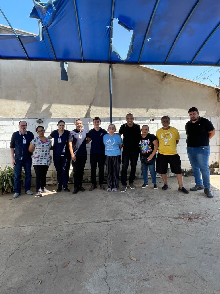 A imagem mostra um grupo de dez pessoas posando para uma foto em frente a uma parede de tijolos brancos. Acima deles, há uma cobertura azul, parcialmente rasgada, com buracos visíveis que deixam passar a luz do sol. As pessoas estão alinhadas lado a lado, com a maioria vestindo roupas casuais. Da esquerda para a direita, vemos: * Um homem com óculos, camisa escura e crachá. * Uma mulher com óculos, blusa estampada e calça escura. * Uma mulher com uniforme escuro e calça escura. * Um homem com camisa escura e calça escura. * Um homem com camisa escura e calça escura. * Uma mulher com uma camiseta azul clara com um símbolo estampado e calça listrada. * Uma mulher com camiseta preta com estampa colorida e calça jeans. * Um homem com uma camiseta amarela com estampa e shorts pretos. * Um homem com camiseta preta e jeans. O chão em frente ao grupo é de concreto, com algumas folhas secas espalhadas. A iluminação sugere que é um dia ensolarado.