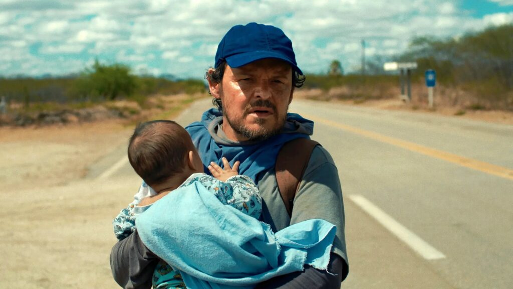 A imagem mostra um homem e um bebê caminhando em uma estrada em um dia ensolarado. O homem usa um boné azul e uma blusa azul. Ele tem barba e parece cansado. Ele segura o bebê em seu braço esquerdo, enrolado em uma manta azul. O bebê está de costas para a câmera. A estrada é pavimentada e tem uma linha amarela no meio. Ao redor da estrada, há terreno seco com alguns arbustos e árvores esparsas. Ao fundo, o céu está azul com algumas nuvens brancas. Há uma placa de sinalização ao lado da estrada à distância. A cena evoca uma sensação de jornada e possivelmente de dificuldades, dada a aparência do homem e o ambiente árido.