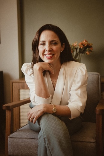 A mulher está sentada em uma poltrona de tecido cinza, com as pernas cruzadas e os braços apoiados no colo. Ela usa uma blusa branca com mangas bufantes e calças verdes. Seu cabelo é escuro e liso, e ela tem um sorriso no rosto, com a mão apoiando o queixo. Ao fundo, há um móvel de madeira com uma cestaria e um vaso com flores alaranjadas. A iluminação é suave e o ambiente parece ser um escritório ou sala de estar aconchegante.