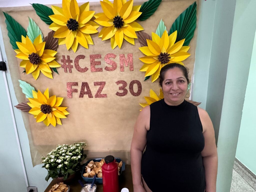 A mulher está sorrindo para a câmera, com um fundo decorado com flores de papel amarelas, verdes e marrons, e letras brilhantes em tom de cobre que dizem "#CESM FAZ 30". Ela está usando um top preto sem mangas e brincos grandes em argola. Na mesa à sua frente, há um buquê de flores brancas, uma travessa com o que parecem ser salgadinhos, uma garrafa de ketchup e outros recipientes com comida.