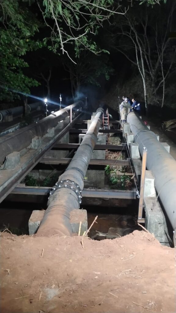 A imagem mostra um trabalho noturno em um grande duto. * *Cenário:* A cena está iluminada por luzes artificiais, indicando que o trabalho está sendo realizado durante a noite. Há árvores e vegetação ao redor, sugerindo um local externo, possivelmente em uma área rural ou industrial com cobertura vegetal. * *Dutos:* Dois grandes dutos de metal são o foco principal. Eles estão elevados do solo e apoiados em estruturas de concreto e metal. Um dos dutos, mais próximo ao espectador, tem uma junta visível com vários parafusos ao redor dela. O outro duto, à direita, parece ser mais liso. * *Trabalhadores:* Duas pessoas vestindo roupas de trabalho estão visíveis perto do duto da direita. Uma delas parece estar em pé, enquanto a outra está mais agachada, possivelmente inspecionando ou trabalhando no duto. Há também uma escada de metal encostada em um dos dutos, indicando acesso para trabalho. * *Atmosfera:* Uma leve fumaça ou vapor pode ser vista saindo de algum ponto próximo ao duto da esquerda, adicionando um elemento dinâmico à cena. O chão ao redor dos dutos parece ser de terra, com algumas áreas escavadas ou em processo de escavação. * *Iluminação:* Várias luzes artificiais estão posicionadas para iluminar a área de trabalho, lançando sombras e destacando os dutos e os trabalhadores.