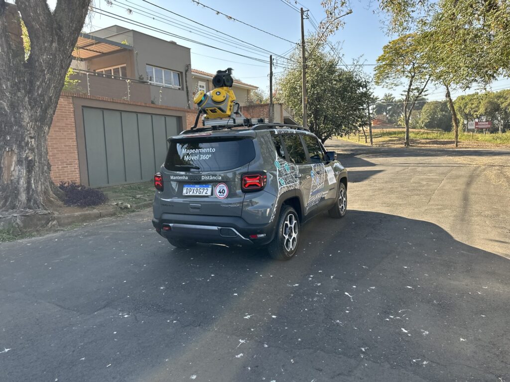 A imagem exibe um Jeep Renegade cinza estacionado em uma rua de asfalto em uma área residencial. O carro está equipado com um dispositivo de mapeamento 360º montado no teto, que é amarelo e preto. Na parte traseira do veículo, há um adesivo com a inscrição "Mapeamento Móvel 360º" e outro logo abaixo dizendo "Mantenha Jeep Distância". A placa do carro é DPX9G72 e há um adesivo de limite de velocidade de 40 km/h ao lado. O lado do carro é decorado com um padrão semelhante a um mapa em tons de verde e branco. Ao lado do carro, à esquerda, há uma árvore grande e uma casa com um portão cinza e uma fachada moderna. Do lado direito, a rua continua em direção a uma área arborizada com uma placa de "VENDE". O céu está claro e azul, indicando um dia ensolarado.