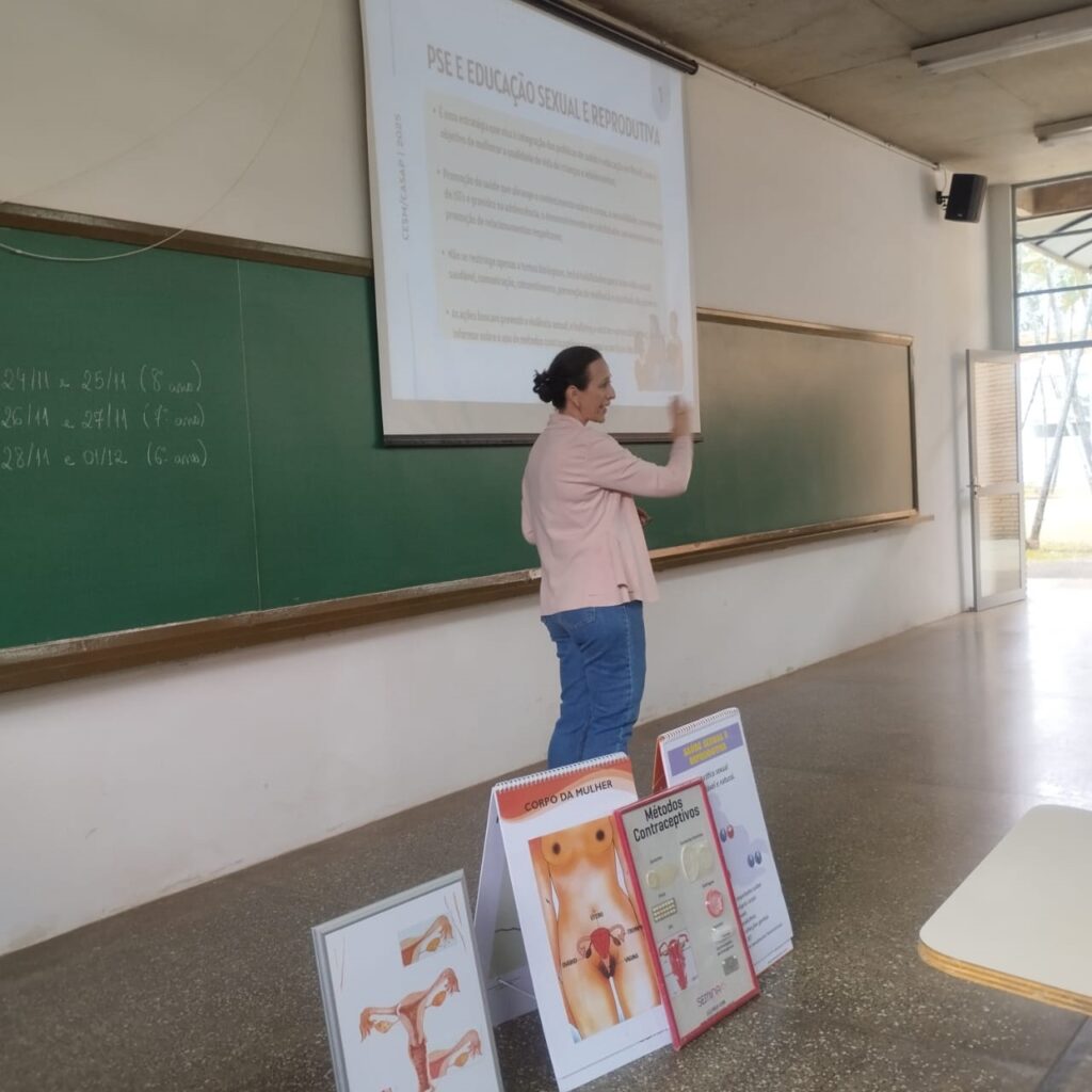 A imagem mostra uma sala de aula onde uma professora está em pé em frente a um quadro negro, apresentando um conteúdo sobre Saúde Sexual e Reprodutiva (PSE). Ela está usando um blazer rosa e calças jeans azuis. Atrás dela, um projetor exibe um slide com o título "PSE E EDUCAÇÃO SEXUAL E REPRODUTIVA" e alguns tópicos listados em português. No quadro negro, à esquerda, há anotações escritas com giz, aparentemente datas e turmas, como "24/11 e 25/11 (8º anos)", "26/11 e 27/11 (7º anos)" e "28/11 e 01/12 (6º anos)". Na frente da área de exposição, há dois expositores gráficos com ilustrações sobre o corpo feminino e métodos contraceptivos. Um deles mostra detalhes do sistema reprodutor feminino com a legenda "CORPO DA MULHER", e outro exibe "Métodos Contraceptivos" com diferentes tipos de pílulas e outros métodos. A disposição dos elementos sugere uma aula expositiva e interativa sobre saúde sexual e reprodutiva, com recursos visuais para auxiliar na compreensão do conteúdo.