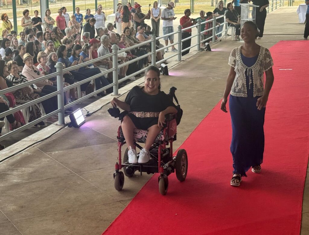 Na imagem, um evento parece estar acontecendo ao ar livre, com um público sentado em arquibancadas à esquerda. No centro, uma mulher em uma cadeira de rodas motorizada, vestindo um vestido preto e tênis brancos, se move em direção à câmera. À direita, uma mulher alta, com um vestido azul longo e um cardigã branco rendado, caminha sobre um tapete vermelho, também em direção à câmera. O cenário sugere uma passarela ou uma cerimônia, com a atenção voltada para as duas mulheres.