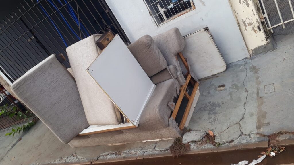 A imagem mostra um sofá velho e danificado, jogado na rua. O sofá está em mau estado, com o tecido rasgado e a estrutura de madeira exposta em algumas partes. Há também um colchão perto do sofá, que parece igualmente desgastado. O cenário sugere que os móveis foram descartados, possivelmente por terem sido substituídos por novos ou por estarem em condições inutilizáveis. O chão ao redor está sujo e rachado, e há alguns detritos espalhados, como folhas secas e pedaços de plástico. Ao fundo, observa-se uma parede com pichações e uma janela com grades, indicando um ambiente urbano.