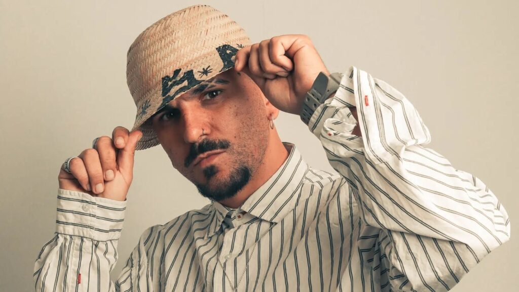 A imagem mostra um close-up de um homem usando um chapéu de palha e uma camisa listrada. Ele está ajustando o chapéu com as mãos, com uma expressão séria no rosto. O homem tem um piercing no nariz, um cavanhaque e usa um anel em um dos dedos. A iluminação é suave e o fundo é de cor clara, destacando o sujeito.