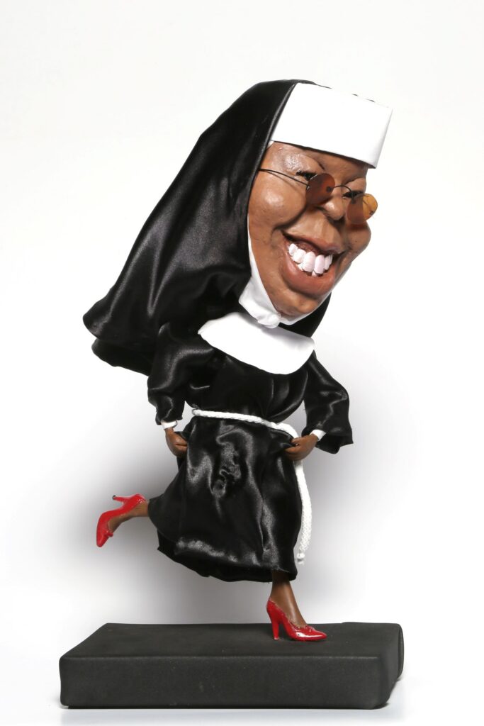 A figura retrata uma caricatura de uma freira em movimento. A personagem está vestida com um hábito preto e branco, característico das freiras, mas com um toque moderno e ousado. O hábito é feito de um tecido preto brilhante, que sugere seda ou cetim, conferindo um ar luxuoso à peça. O capuz e o véu são pretos e cobrem a cabeça e parte do rosto, como é tradicional. Um colarín branco, também conhecido como "corpo" ou "jabot", destaca-se no pescoço, em contraste com o preto do hábito. O que mais chama a atenção na figura são os detalhes que fogem do tradicional. A freira usa óculos de sol com lentes arredondadas e armação fina, que lhe dão um ar descontraído e jovial. Seu rosto é caricaturado, com um sorriso largo e dentes aparentes, transmitindo alegria e vivacidade. A pele da figura tem um tom escuro, característica da atriz Whoopi Goldberg, que interpretou uma freira no filme "Mudança de Hábito". A figura está em movimento, com uma perna levantada e a outra apoiada em um pedestal preto. Ela usa sapatos de salto alto vermelhos, um elemento completamente inesperado e ousado para uma freira, que adiciona um toque de glamour e irreverência à imagem. O vestido preto é ajustado na cintura por um cordão branco trançado, que também foge do visual mais austero das vestimentas religiosas convencionais. A base onde a figura está apoiada é um bloco retangular de cor preta, simples e discreto, que serve para destacar a personagem em si. O fundo da imagem é branco, o que realça ainda mais as cores e os detalhes da figura. A iluminação é frontal e uniforme, permitindo a visualização clara de todos os elementos.