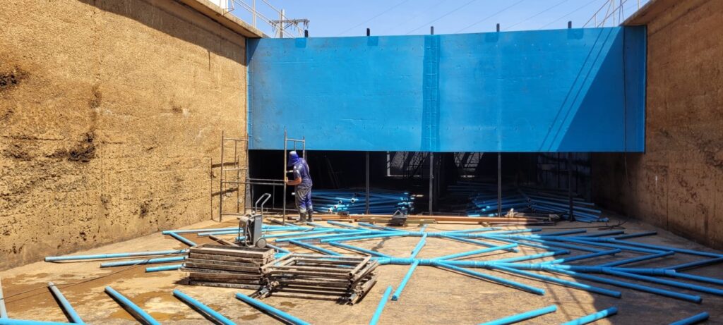 A imagem mostra uma cena de construção dentro de um grande tanque ou reservatório com paredes de concreto. Vários trabalhadores estão ocupados com a montagem de estruturas metálicas no chão. * A maioria dos trabalhadores está vestida com uniformes azuis e usa chapéus para se proteger do sol ou de detritos. * A estrutura metálica parece ser composta por longas vigas pretas fixadas ao chão por suportes em forma de L. * Há pilhas de materiais de construção, incluindo metal e madeira, espalhadas pela área. * Equipamentos como andaimes e possivelmente ferramentas elétricas também são visíveis. * O chão do reservatório é de concreto, com algumas áreas molhadas ou com manchas de água. * As paredes do reservatório são de cor ocre, com textura áspera e algumas marcas de umidade. * A iluminação sugere um dia claro, possivelmente ensolarado, com sombras projetadas pelos trabalhadores e pelas estruturas.